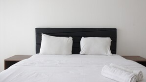 A neatly made bed with white linens and pillows, a dark headboard, and a wooden nightstand beside it. - Spacious Studio Room at Galeri Ciumbuleuit 3 Apartment (Bandung)