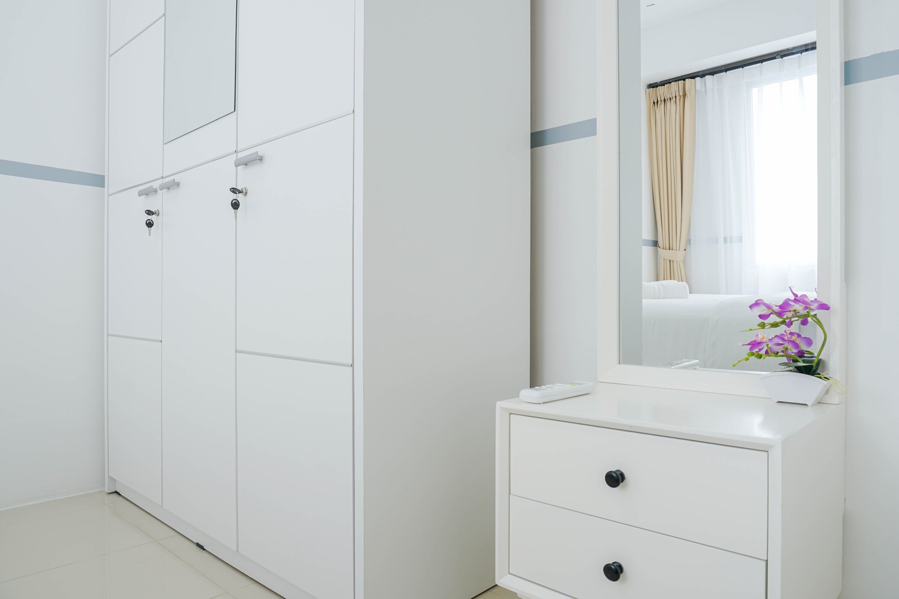 A white wardrobe with multiple compartments, a mirror, and a small bedside table with a vase of flowers.