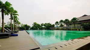 A swimming pool with a wooden deck and lounge chairs, surrounded by palm trees and a building with a tiled roof. - Best Location and New Furnished @ Studio Capitol Park Residence Apartment (Jakarta)