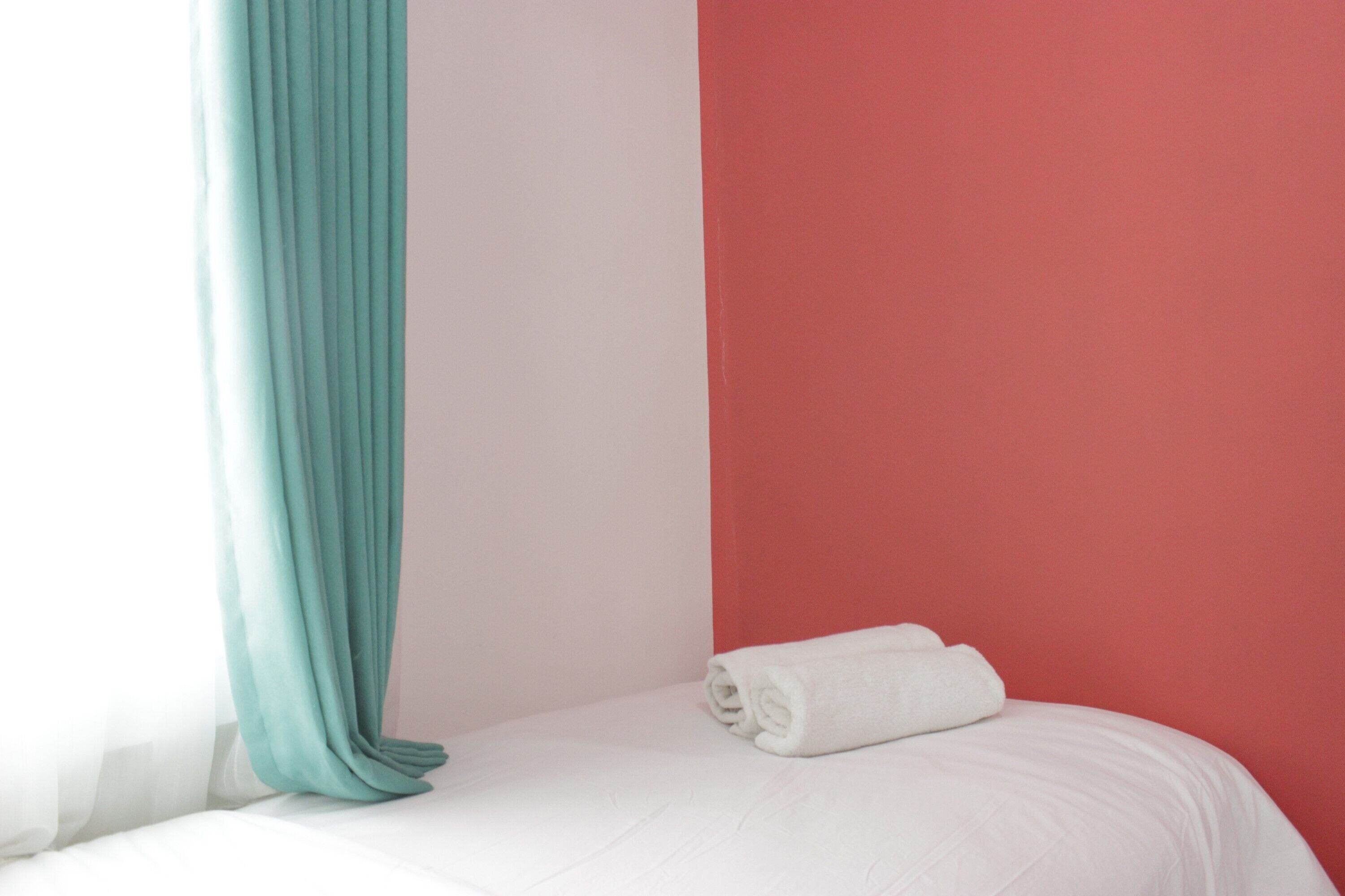 Un lit bien fait, avec un drap blanc et une serviette blanche enroulée, un rideau turquoise et un mur rouge.