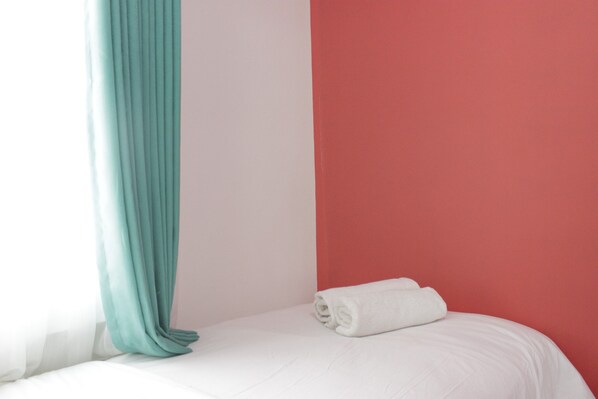 Una cama bien hecha con sábana blanca y una toalla blanca enrollada, una cortina color turquesa y una pared roja.