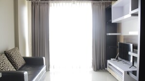 A modern living room with a black sofa, patterned cushions, a flat-screen TV on a white entertainment unit, and a large window with grey curtains. - Spacious 2BR Apartment Gateway Pasteur near Exit Toll 23 (Cimahi)
