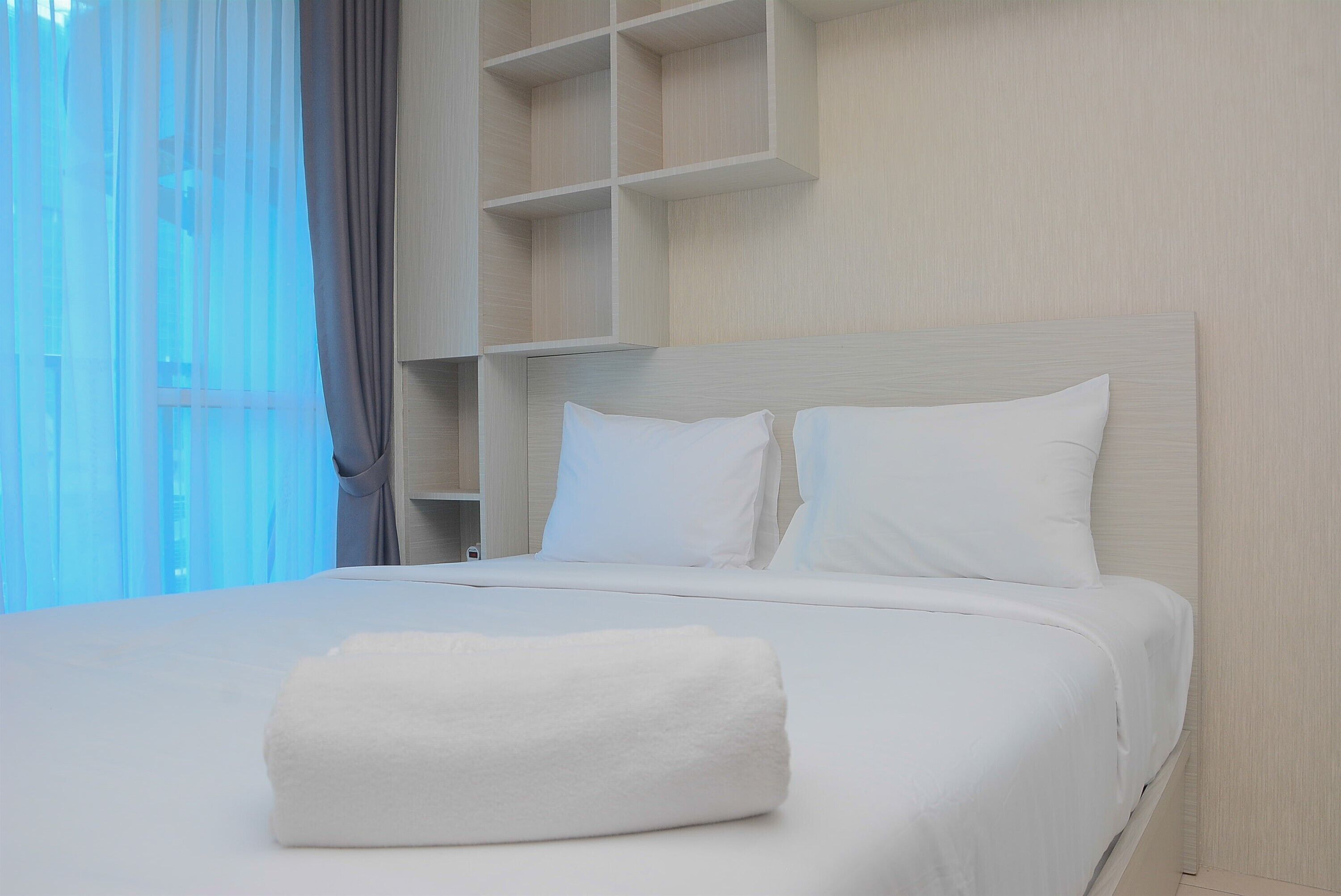 A neatly made bed with white linens and pillows, a rolled white towel on the bed, and a light-colored wall with a built-in shelving unit.