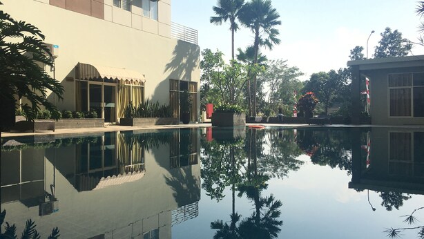 A swimming pool with clear reflections, surrounded by buildings and palm trees. - Chic and Cozy 2BR Apartment at Pinewood Jatinangor near JATOS (Cileunyi)