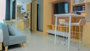 A modern living room with a sofa, a dining area with stools, a TV mounted on the wall, and a kitchen area with a microwave. - Grand Kamala Lagoon Studio Apartment with Modern Interior (Bekasi)