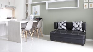 A modern kitchen with white cabinets, a dining area with a white table and chairs, and a black sofa with patterned cushions. - Spacious 3BR Apartment Parahyangan Residence near UNPAR (Bandung)