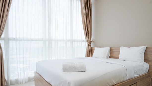 A neatly made bed with white linens and a folded towel, wooden headboard, and sheer curtains. - Cozy and Wonderful 1BR Ciputra International Apartment (Jakarta)
