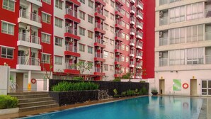 A swimming pool with a tiled edge, surrounded by a building with red and white exterior walls, balconies, and a stone wall with plants.