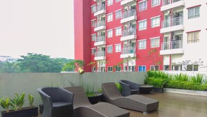 A rooftop lounge with wicker furniture, potted plants, and a red building in the background.