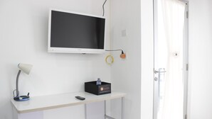 A room with a white desk, a flat-screen TV mounted on the wall, a lamp, a remote control, and a travel bag on the desk.