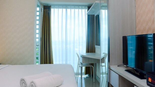 A hotel room with a bed, a desk, a chair, a television, and a window with curtains. - Comfy with Modern Style 1BR Grand Kamala Lagoon Apartment (Bekasi)