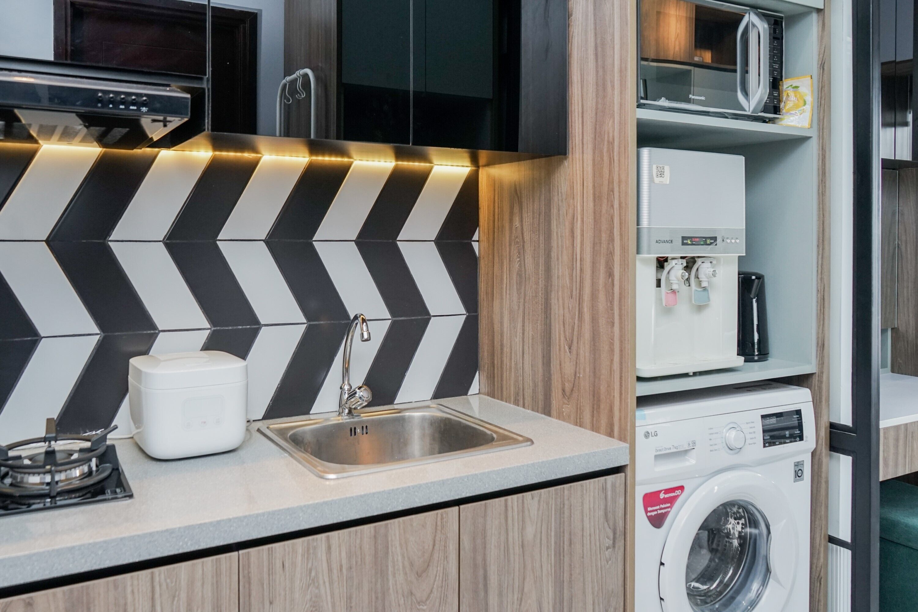 A compact kitchen with a modern design, featuring a black and white chevron backsplash, a stainless steel sink, a built-in microwave, and a front-loading washing machine.