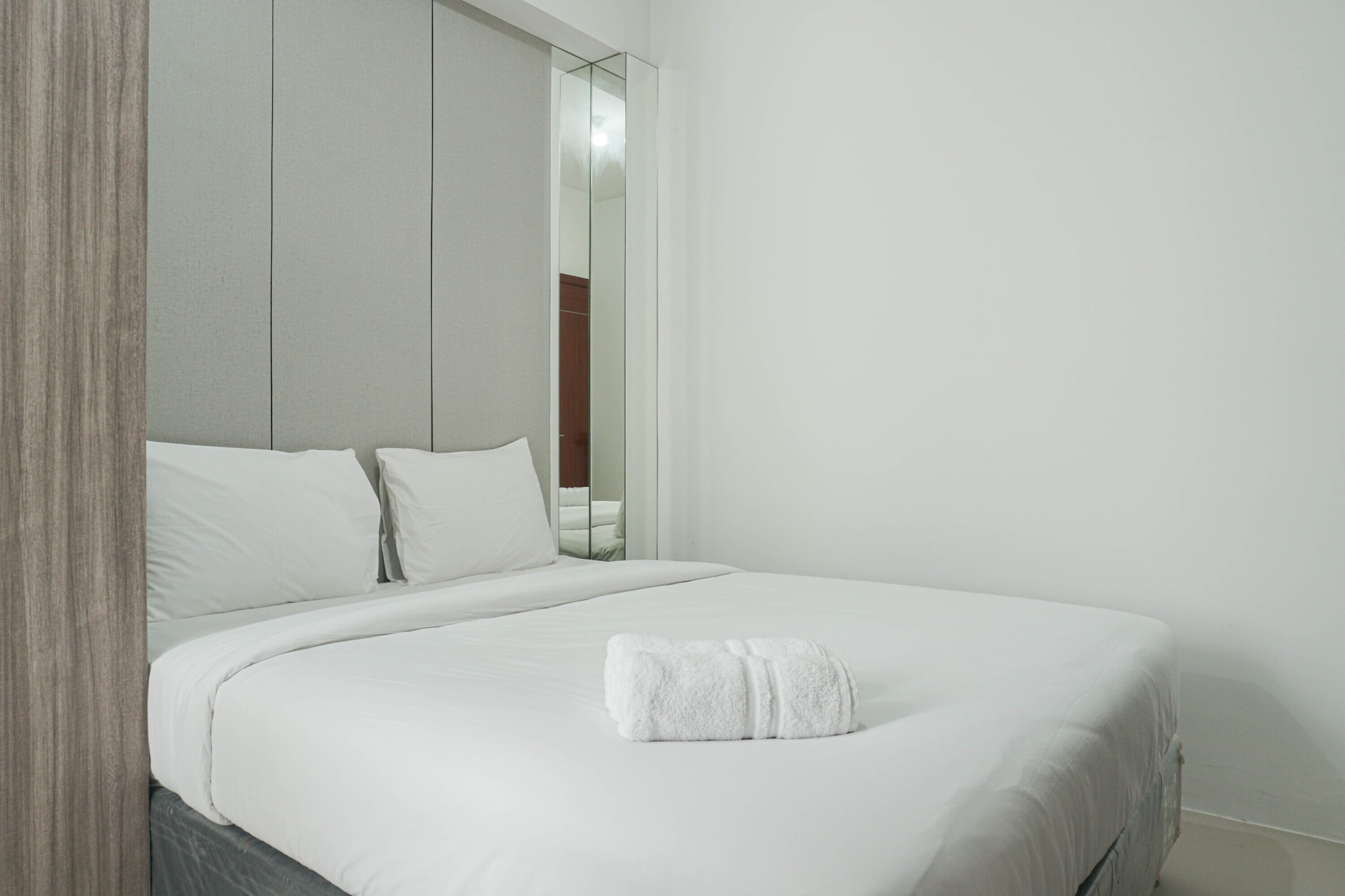A neatly made bed with white linens and a rolled white towel in a modern, minimalist room with a wooden headboard and a large mirror.