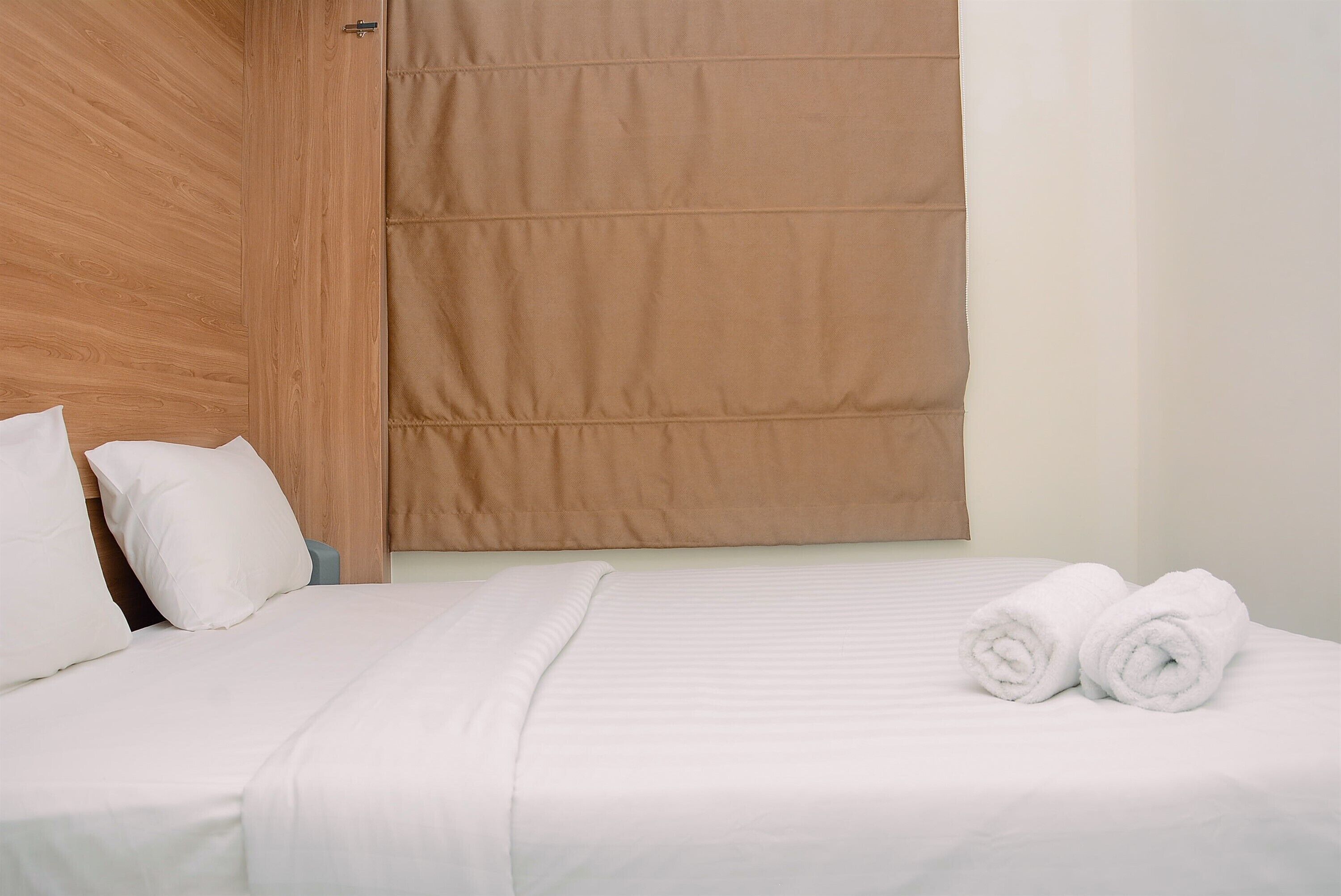 A neatly made bed with white linens and two rolled towels, a wooden headboard, and a beige curtain.