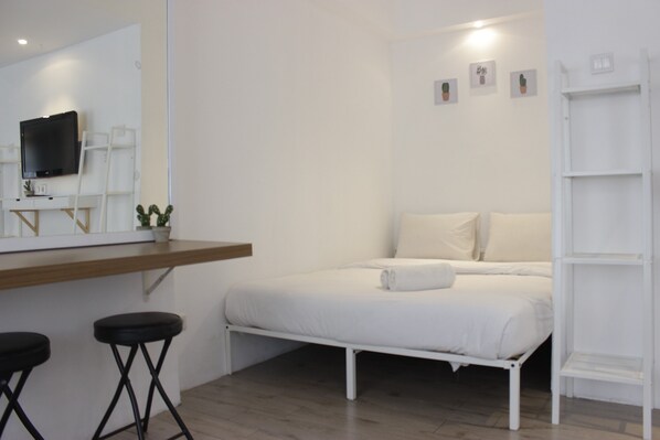 A minimalist bedroom with a bed, a desk with a flat-screen TV, and a small plant.