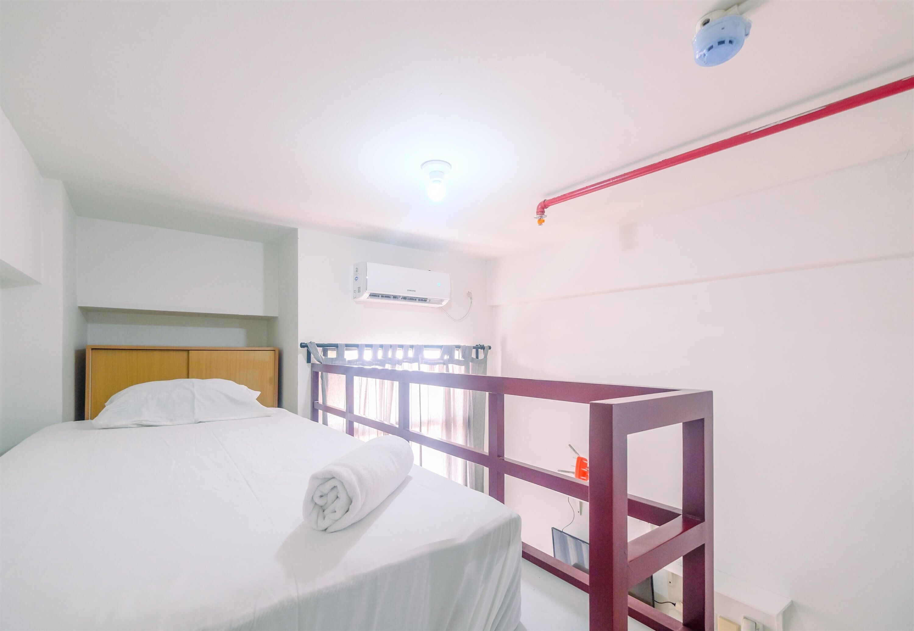 A single bed with a wooden headboard, a white blanket, and a rolled towel. There is a red-framed bunk bed to the right. An air conditioning unit is mounted on the wall above the bed.