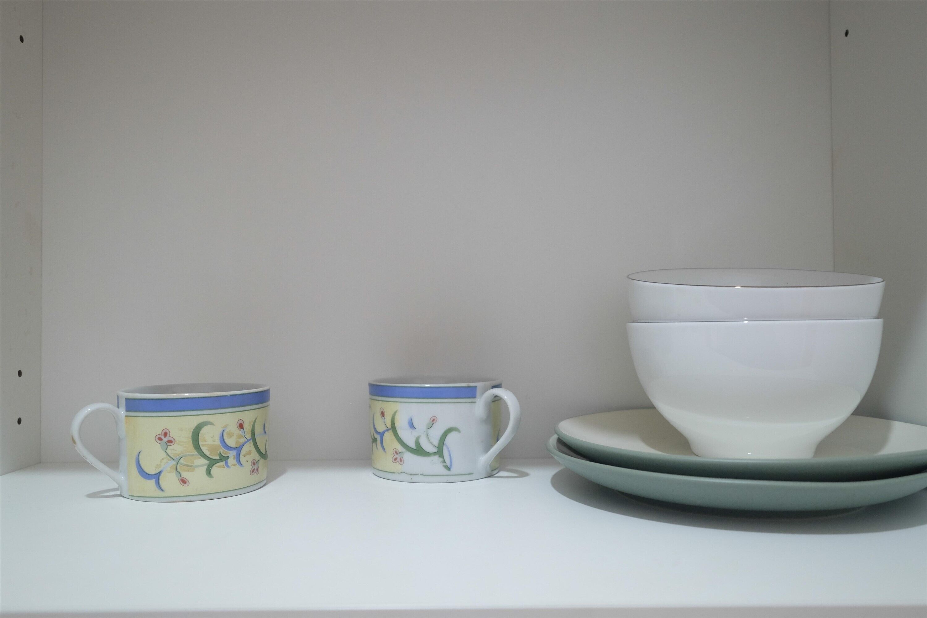 Una estantería blanca con una taza decorativa, una taza más pequeña y un bol blanco sobre un plato a juego.
