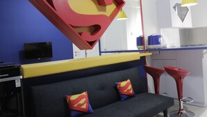 A modern room with a sofa, a large Superman logo decoration, a coffee machine, and a bar area with stools. - Fabolous Thematic 2BR Apartment at Parahyangan Residence Near UNPAR (Bandung)