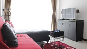 Une pièce avec un canapé rouge, un pouf noir, un téléviseur posé sur un meuble et une petite table surmontée d’un tapis rouge.