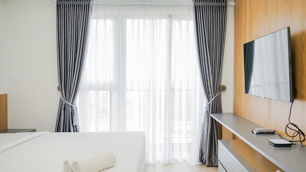 Ein Hotelzimmer mit Bett, Fernseher an einer Holzvertäfelung, Schreibtisch mit Laptop und Fenster mit durchsichtigen Vorhängen.