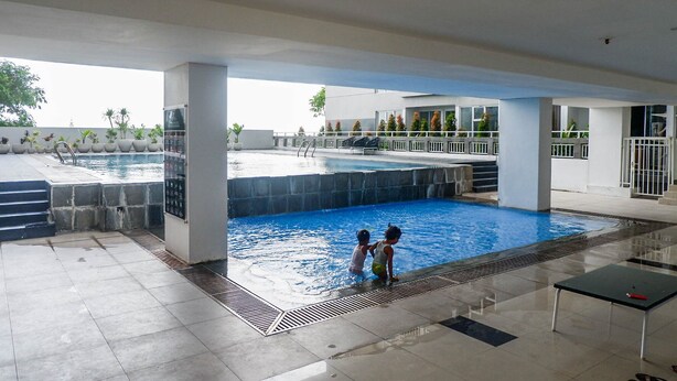 Ein Schwimmbeckenbereich mit zwei spielenden Kindern, umgeben von einer überdachten Terrasse und einem Gebäude mit weißer Fassade.