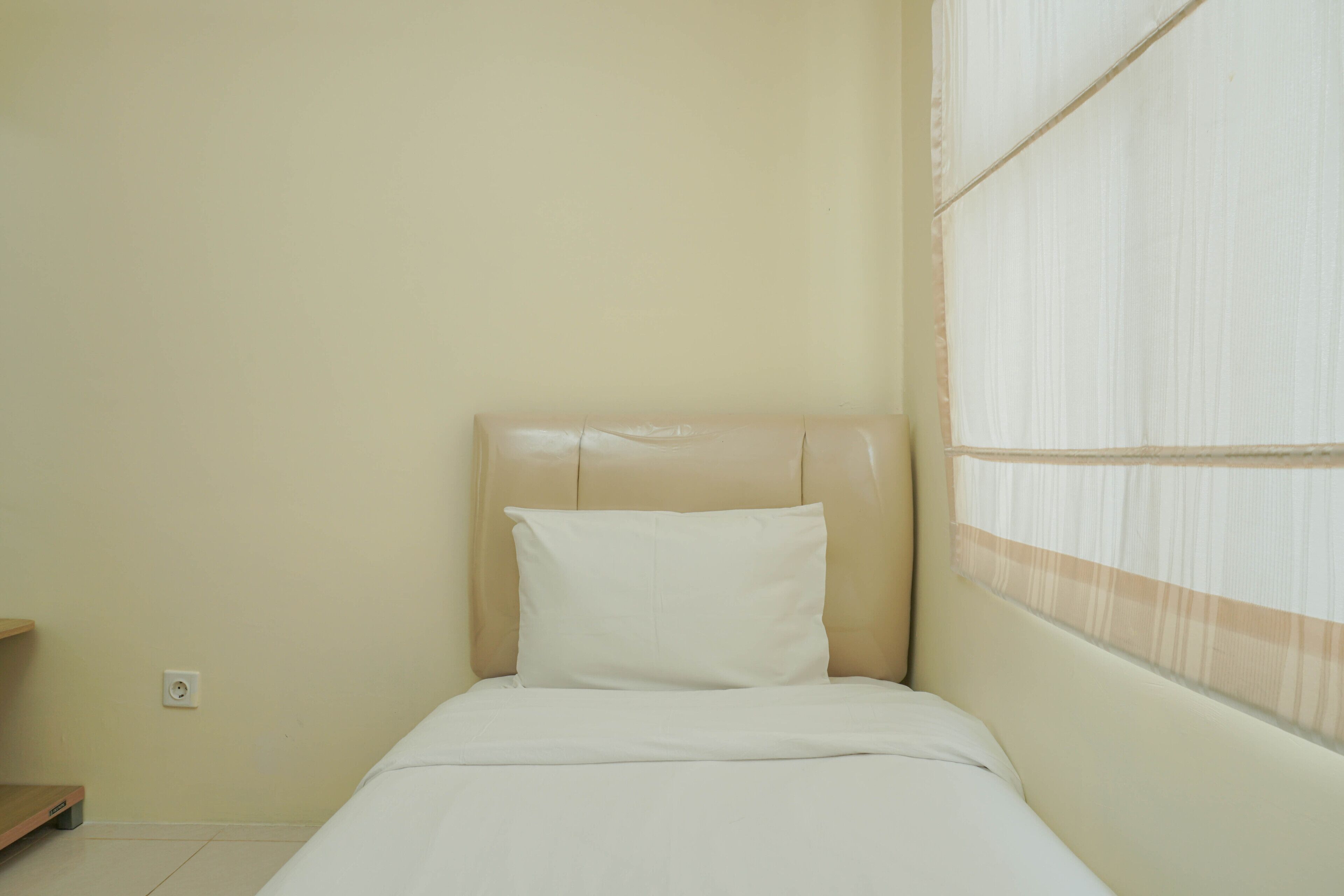 A small, neatly made bed with a headboard and a window with sheer curtains.