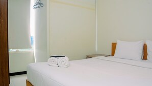 Een net opgemaakt bed met witte lakens en twee opgerolde handdoeken, een houten hoofdbord en een klein houten nachtkastje met een lamp.