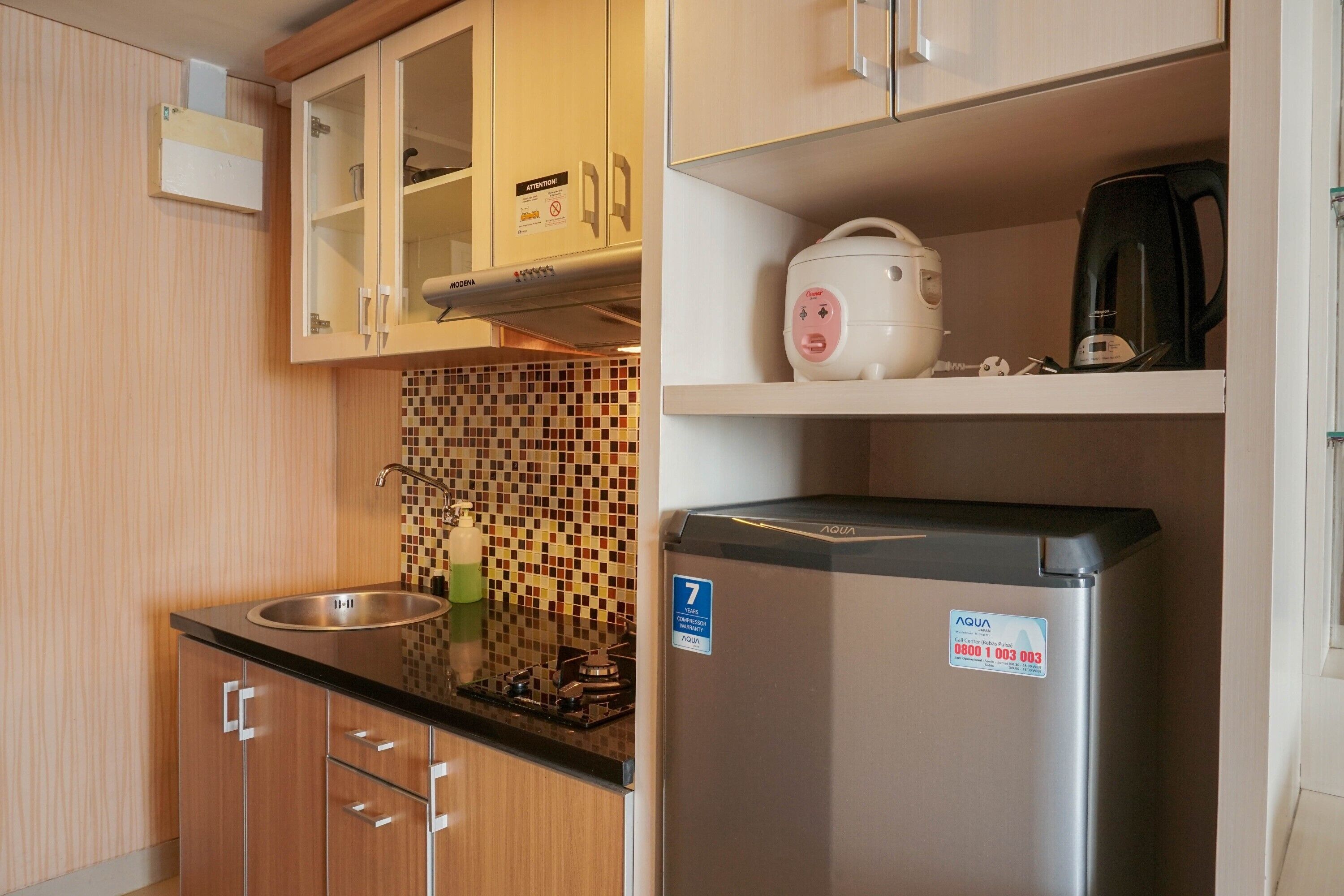 A compact kitchen with wooden cabinets, a stainless steel sink, a tiled backsplash, and a refrigerator with a water dispenser.