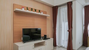 A modern living room with a flat-screen TV on a white cabinet, a shelf with decorative items above, and brown curtains on the window. - Elegant and Relaxing Studio Apartment H Residence (Jakarta)