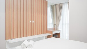A modern hotel room with a large bed, a wooden panel wall, a small desk, and a window with curtains. - Marvelous 1BR at The Branz Apartment (Pagedangan)