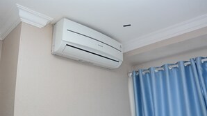 A white air conditioning unit mounted on a wall in a room with a blue curtain. - Spacious and Comfy 2BR Bassura City Apartment (Jakarta)