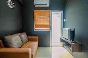 Ein modernes Wohnzimmer mit einer Ledersofa, einem Flachbildfernseher auf einem Holzständer, einer Wandklimaanlage und einem Fenster mit Jalousien.