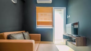 A modern living room with a sofa, a television, and a window with blinds. - Comfort 1BR with Study Room Green Pramuka Apartment (Jakarta)