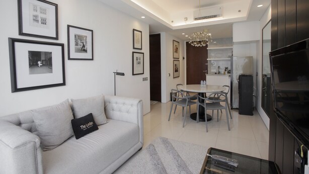 A modern living room with a sofa, framed pictures, a dining area with a table and chairs, and a kitchen in the background. - Premium 2BR Apartment near Marvell City Mall at The Linden (Surabaya)