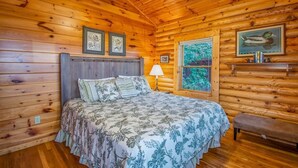 Un dormitorio tipo cabaña de troncos con cama, un banco y una ventana con vistas a follaje.