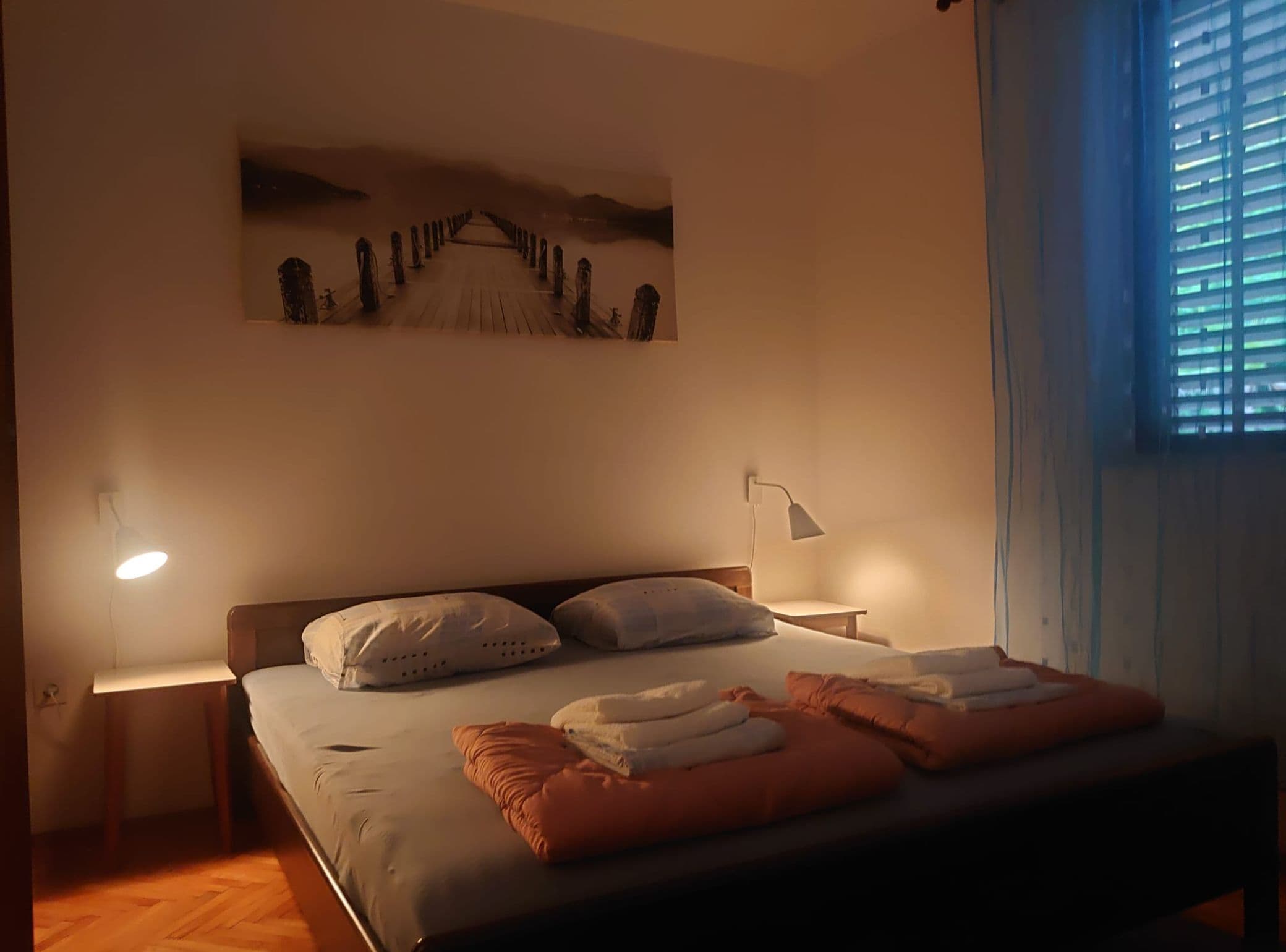 A bedroom with a bed, a nightstand, a wall-mounted lamp, and a window with blinds.