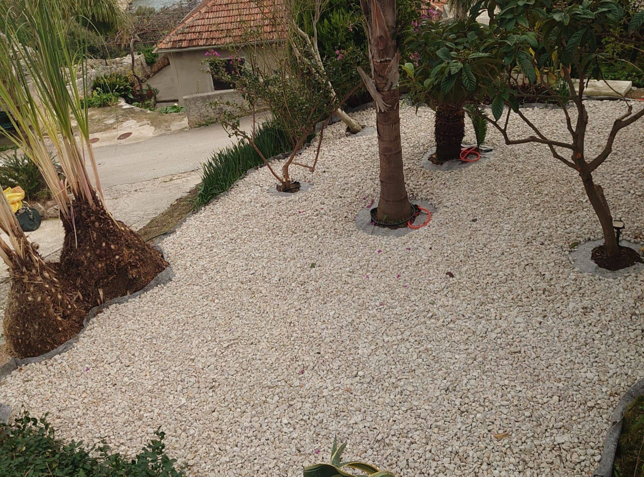 Een tuin met grindpaden, diverse beplanting en een klein gebouw met pannendak.