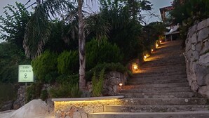 Una escalera de piedra conduce a la entrada de una villa, rodeada de abundante vegetación y un letrero que da la bienvenida a Villa Peragic.