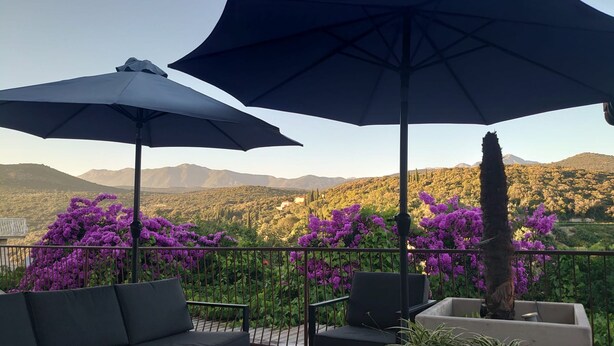 Una terraza con mobiliario exterior, incluyendo un sofá y sillas, bajo grandes sombrillas, con vistas a montañas y flores púrpuras al fondo.