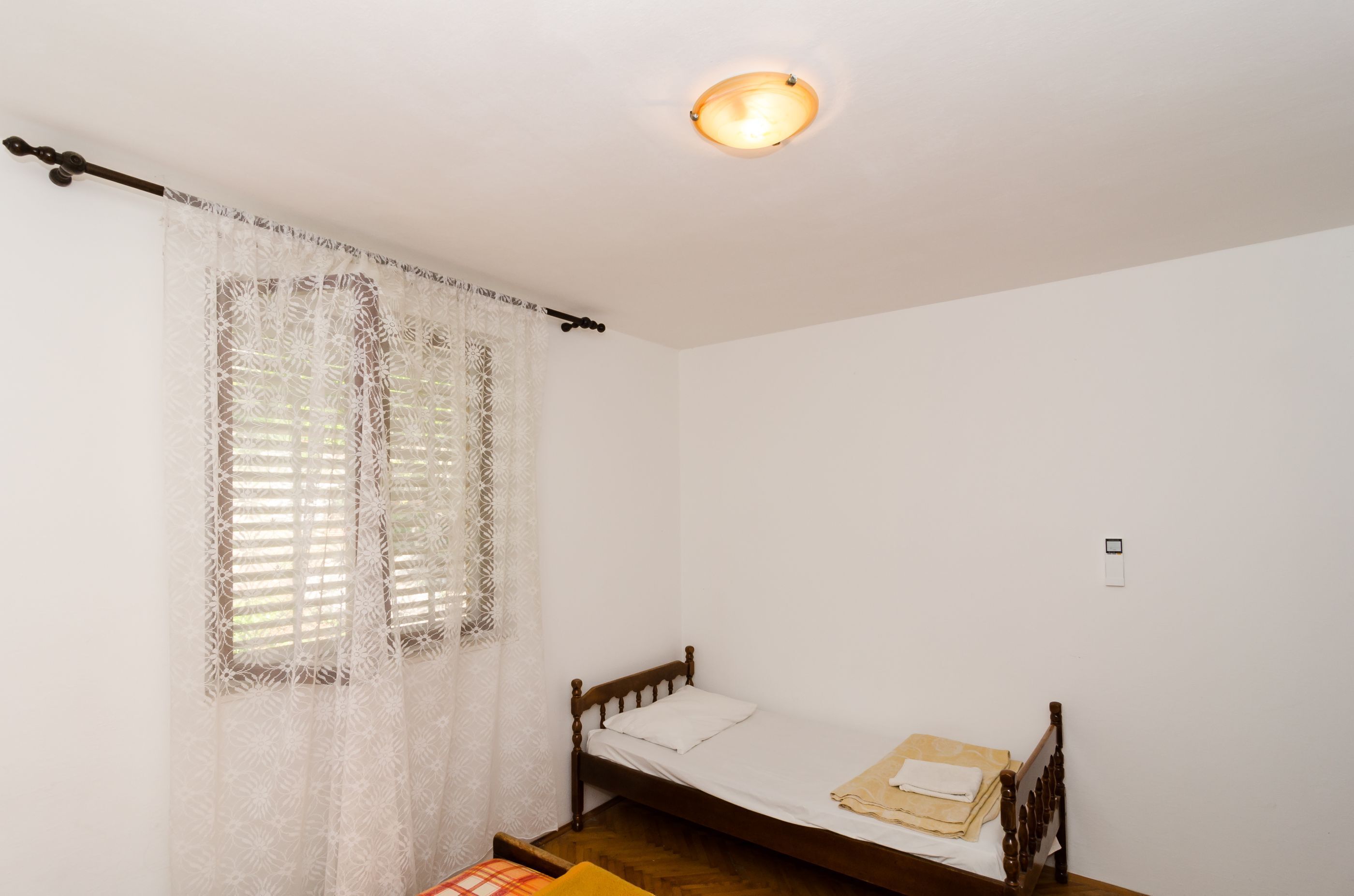 A small, single-bed room with a wooden headboard, a white bedspread, and a window with lace curtains.