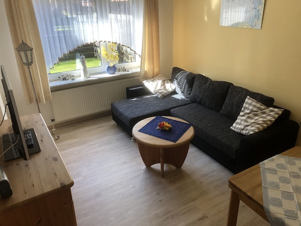 Uma sala de estar com um sofá cinza escuro, uma mesa de centro redonda de madeira com um jogo americano azul, um radiador, uma janela com cortinas leves e vista para um jardim.