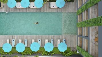 A rooftop pool area with multiple sun loungers and umbrellas, surrounded by lush greenery and a wooden deck.