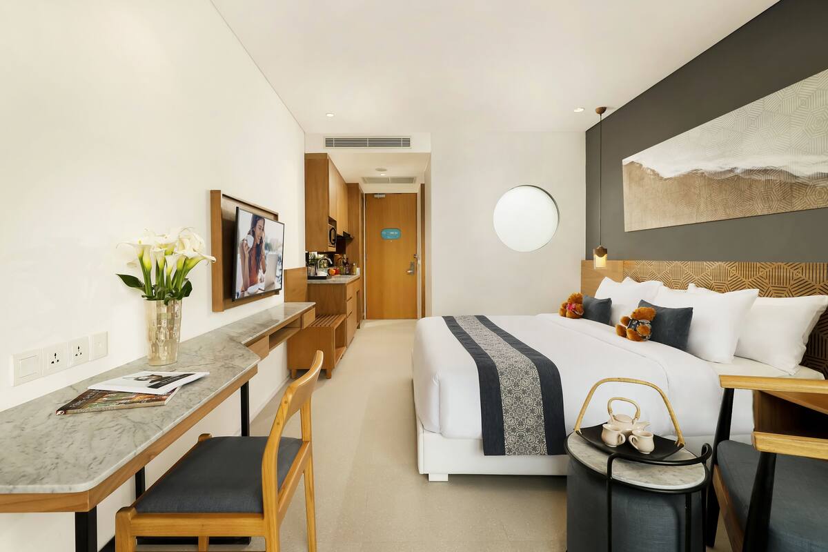 Habitación de hotel con cama, escritorio, silla, una mesita con tetera, un florero con flores y un cuadro en la pared.