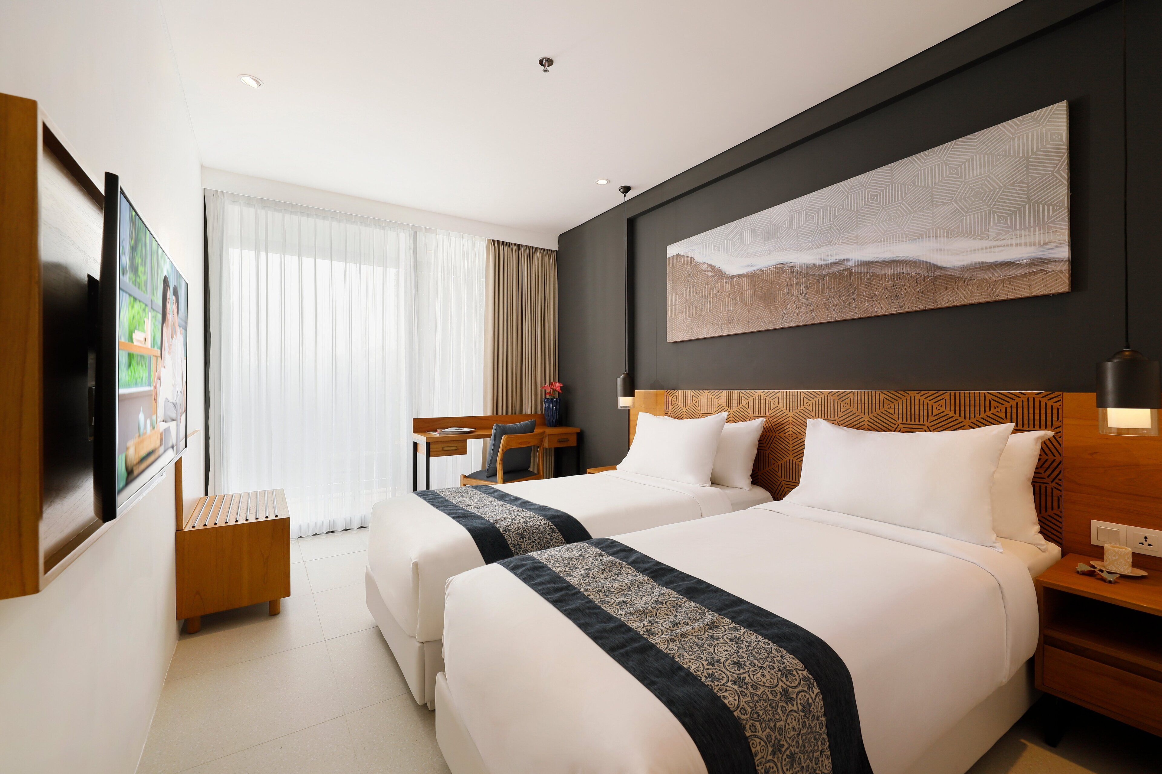 Ein Hotelzimmer mit zwei Betten, einem Schreibtisch, einem Stuhl, einem Fernseher und einem großen Fenster mit Vorhängen.