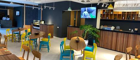 A modern restaurant interior with wooden tables and chairs, a bar area, and a television screen displaying a program.