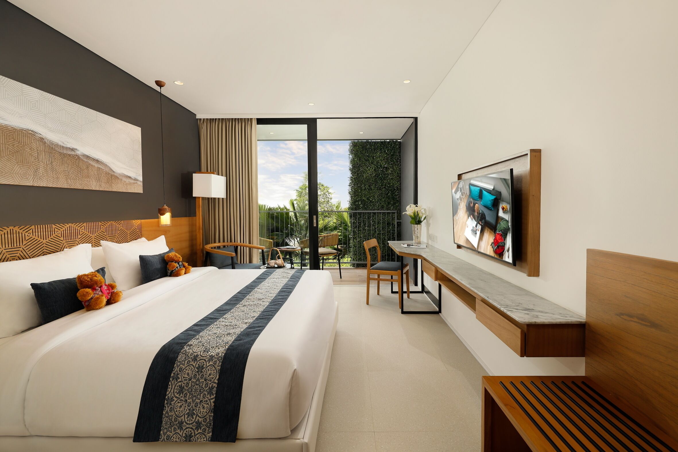A modern hotel room with a large bed, a desk, a chair, a TV, and a balcony with a view of greenery.
