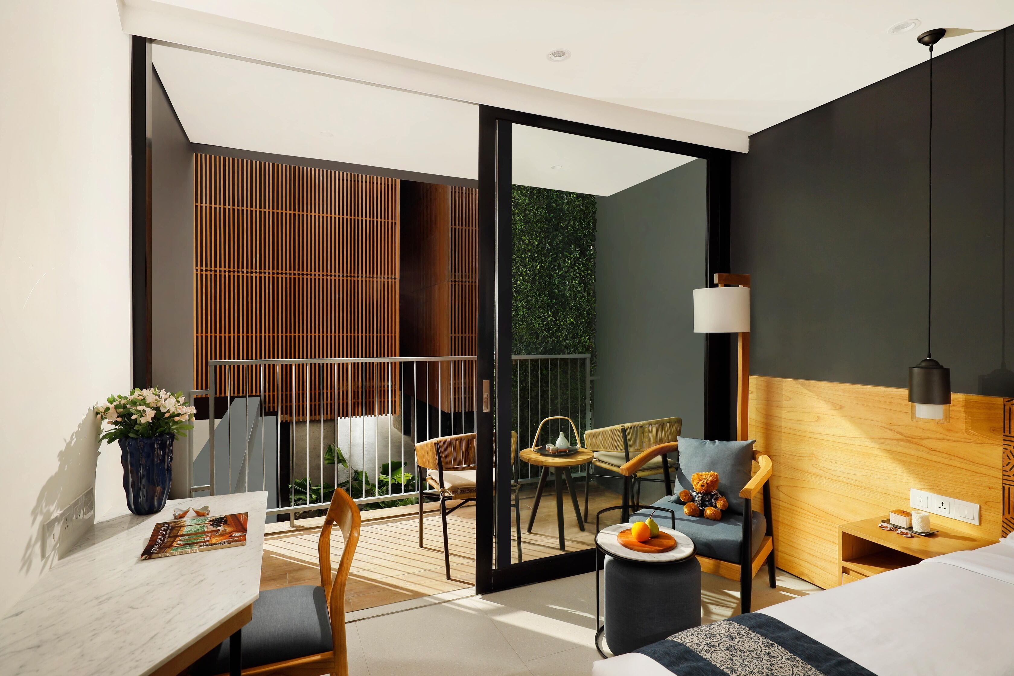 A modern hotel room with a large bed, a dining table with chairs, a small seating area with a sofa and chairs, a wooden side table with a lamp, and a view of a balcony with plants and a railing.