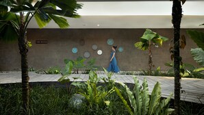 Una mujer con vestido azul camina en un jardín interior moderno con plantas tropicales y una pared decorada con diseños circulares.