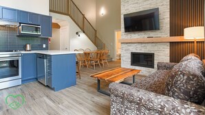 A modern kitchen with blue cabinets, a microwave, and a coffee pot. There is a dining area with wooden chairs and a table. A staircase leads to an upper level. A TV is mounted on the wall above a fireplace. - 2 BDR w Loft / Incredible Views / Family-Friendly (Fernie)
