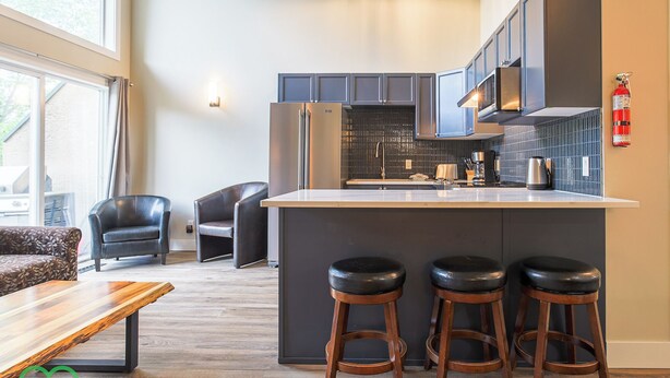 A modern kitchen with dark cabinetry, a central island with stools, and a view of the outdoors. - 2 BDR w Loft / Incredible Views / Family-Friendly (Fernie)
